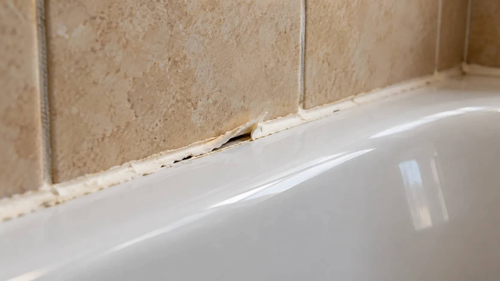 Peeling white caulk separating from the joint between a bathtub and tiled wall